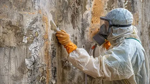 Remediation technician in a respirator scraping mold from a damp wall